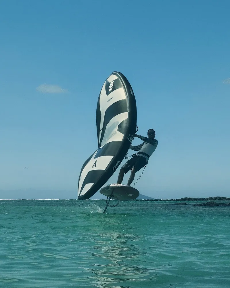 Ludovic wingfoiling above a turquoise lagoon in Mauritius, wing in hand, board flying on its foil